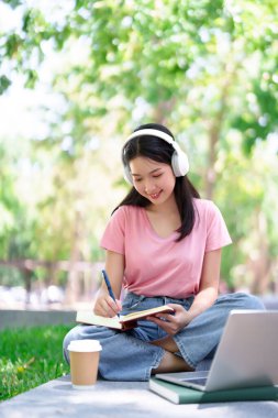 Asyalı genç kız öğrenci eğitim konsepti olarak dizüstü bilgisayar kulaklıkları ve kahve ile parkta öğrenmeyi seviyor. Uzak çalışma çevrimiçi öğrenim yaşam tarzı.