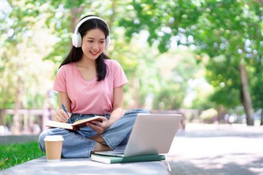 Genç Asyalı bir kadın, dizüstü bilgisayar kulaklıklarıyla parkta ders çalışıyor. Gülümsüyor, notlar alıyor ve dışarıdaki öğrenme ortamının tadını çıkarıyor. Eğitim ve çevrimiçi kurslar için mükemmel..