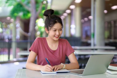 Genç bir Asyalı kadın online çalışmalarına dizüstü bilgisayar kullanarak ve not alarak odaklanıyor. Bu görüntü modern eğitimin ve akademik başarının özünü yansıtır..