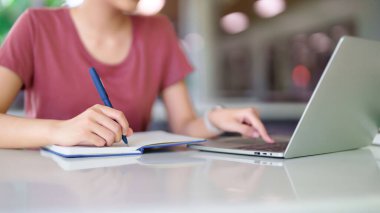 Genç Asyalı kadın dizüstü bilgisayarla ders çalışıyor, not defterine notlar yazıyor. Yeteneklerini geliştirmek için çevrimiçi kursa gidiyor. Eğitim, teknoloji, öğrenme kavramı.