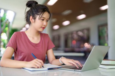 Genç Asyalı bir kadın dizüstü bilgisayarda çalışıyor ve bir deftere yazı yazıyor. İnternette konsantre öğrenci öğrenimi, kütüphanede ya da çalışma alanında notlar almak. Eğitim kavramı.