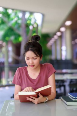 Genç Asyalı bir kadın bir kitap okumaya, muhtemelen bir sınava hazırlanmaya, kütüphanedeki masada oturmaya, bilgi ve eğitimin artmasına odaklanıyor..
