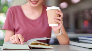 Genç Asyalı kadın çalışırken kahve molasından zevk alır. Masada kitap okuyor. Kolej kampüsü ortamında rahatlamak ve dinlenmek için zaman ayırıyor..