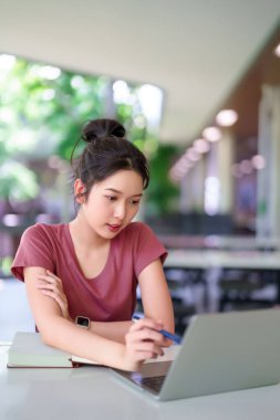 Genç bir Asyalı kadın kütüphane ortamında dizüstü bilgisayar kullanarak dikkatle çalışıyor. Çevrimiçi eğitime odaklanmış, çalışkan öğrenim alışkanlıkları ile becerileri geliştirerek akademik başarıyı temsil etmektedir..