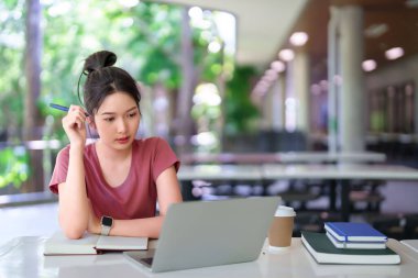 Asyalı kız öğrenci kütüphanede online ders için dizüstü bilgisayar kullanıyor ve not defterine notlar yazıyor. Eğitime odaklanmış. Bilgi ve sınav hazırlığı için üniversite eğitimi.