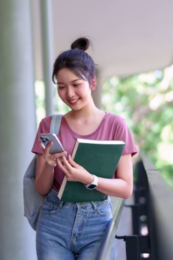 Genç bir Asyalı öğrenci kampüste elinde kitap tutarken akıllı telefonunu kullanıyor. Modern eğitim yolculuğu ve akademik hayatı somutlaştırıyor..