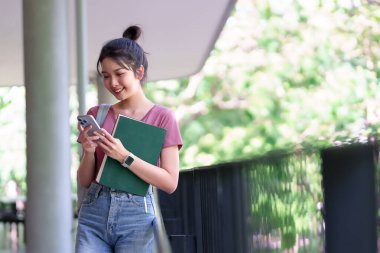 Genç bir Asyalı kadın akıllı telefonunu kullanarak gülümsüyor ve bir üniversite kampüsünde kitap tutuyor. Eğitim, üniversite, lise veya içerik eğitimi için mükemmel.