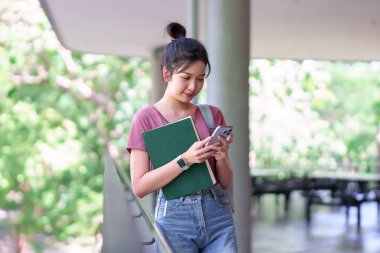 Üniversite kampüsünde okurken akıllı telefon kullanan genç bir Asyalı kız öğrenci. O bir kitap tutuyor ve gülümsüyor. Eğitim konsepti, açık hava sahnesi.