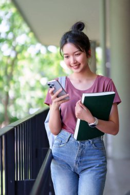 Kampüste cep telefonu kullanan Asyalı genç bir kadın. Mutlu kız öğrenci mesajlaşması, eğitim araştırması. Üniversite bilgisi için okula dönüyorum..