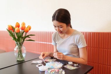 Konforlu ev masasında rahatlatmak için el yapımı boncuklu mücevherler yapan genç bir kadın.