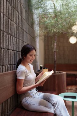 Kadın, doğal ışık ve ahşap dekorlu huzurlu bir kafede kitap okurken kahveden zevk alıyor.