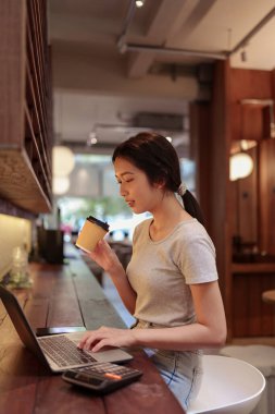 Kahve içen genç bir kadın, şık bir kafe tezgahında dizüstü bilgisayarla çalışırken sıcak ışıklandırmalı.