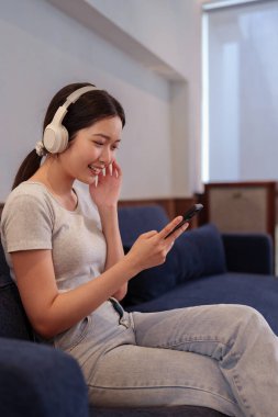 Kanepede oturup kulaklıkla müzik dinleyen ve akıllı telefondan şarkı okuyan genç bir kadın.