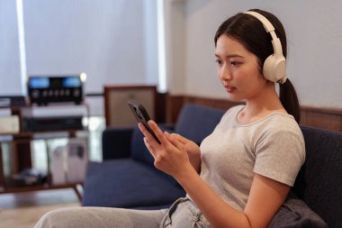 Kanepede oturup kulaklıkla müzik dinleyen ve akıllı telefondan şarkı okuyan genç bir kadın.