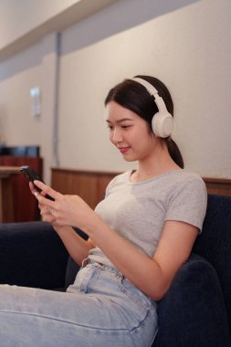 Kanepede oturup kulaklıkla müzik dinleyen ve akıllı telefondan şarkı okuyan genç bir kadın.