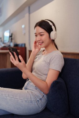 Kanepede oturup kulaklıkla müzik dinleyen ve akıllı telefondan şarkı okuyan genç bir kadın.