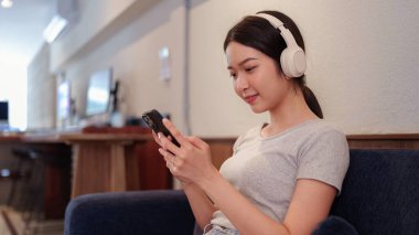 Kanepede oturup kulaklıkla müzik dinleyen ve akıllı telefondan şarkı okuyan genç bir kadın.