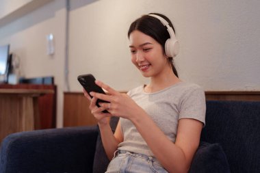 Kanepede oturup kulaklıkla müzik dinleyen ve akıllı telefondan şarkı okuyan genç bir kadın.