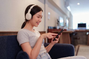 Kanepede oturup kulaklıkla müzik dinleyen ve akıllı telefondan şarkı okuyan genç bir kadın.