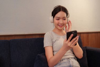 Kanepede oturup kulaklıkla müzik dinleyen ve akıllı telefondan şarkı okuyan genç bir kadın.