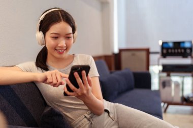 Kanepede oturup kulaklıkla müzik dinleyen ve akıllı telefondan şarkı okuyan genç bir kadın.