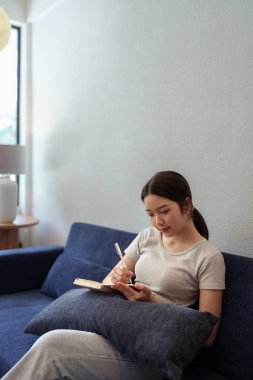 Sakin bir genç kadın rahat bir yuva ortamında kanepede oturup günlüğünü yazıyor.