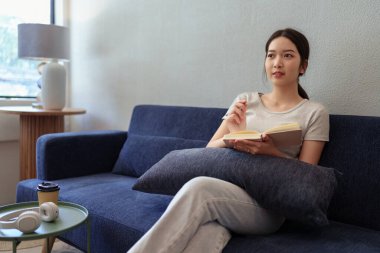 Sakin bir genç kadın rahat bir yuva ortamında kanepede oturup günlüğünü yazıyor.