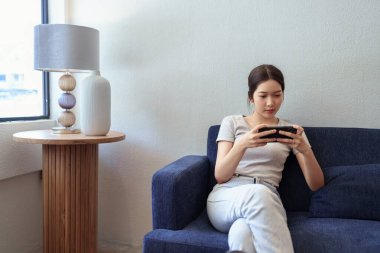 Gündüz vakti rahat bir kafede kanepede otururken akıllı telefondan eğlenen genç bir kadın.