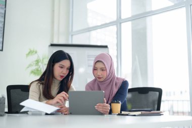 İki genç Asyalı kadın, biri tesettür takıyor, modern bir ofis masasında bir tablet üzerinde iş fikirleri ve stratejilerini tartışıyor. Müslüman iş kavramı.