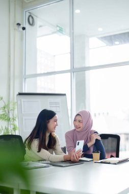 İki genç Asyalı kadın, biri tesettür takıyor, modern bir ofiste işbirliği yapıyorlar, akıllı telefon kullanarak iş çözümleri ve onların Müslüman işleri için fikirleri tartışıyorlar..