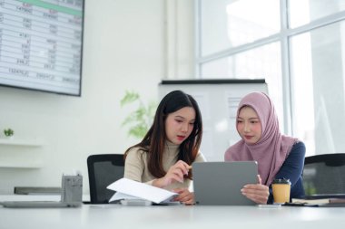 Biri tesettürlü iki genç Asyalı kadın, modern bir ofiste dijital tablet kullanarak Müslüman bir iş kurma girişiminde işbirliği yapıyorlar. Profesyonel takım çalışmasının vurgulanması & başarılı