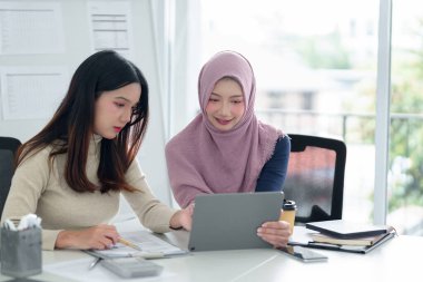 İki genç Asyalı kadın, biri tesettürlü, iş analizi için tablet kullanıyorlar. Parlak, modern bir ofiste finansal raporları tartışıyoruz. Girişim işi takım çalışması kavramı.