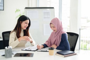 İki genç Asyalı kadın girişimci, biri tesettür takıyor, Müslüman iş stratejisinde işbirliği yapıyor, modern bir ofis çalışma alanındaki belgeleri inceliyor.