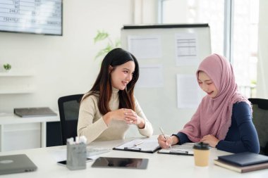 İki Asyalı kadın, biri tesettür takıyor, iş toplantısında planlarını tartışıyorlar. Bunlar Müslüman ticareti, İslami finans ve girişimciliği temsil ediyor. Bu başarı için bir işbirliği..