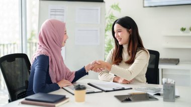 İki genç Asyalı kadın, biri tesettür takıyor, iş toplantısında el sıkışıyor. Müslüman ticaretinde başarılı bir anlaşmayı, ortaklığı ve profesyonel işbirliğini simgeleyen modern bir ofisteler..