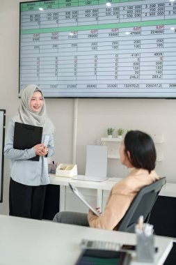 Ofiste iki iş kadını bir ekranda finansal verileri analiz ediyor. Tesettürlü Asyalı bir kadın meslektaşına veri sunar. İş büyümesi için eğilimler, satışlar, kârlar ve stratejiler tartışılıyor.