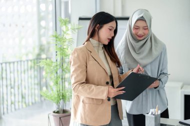 Biri tesettürlü iki Asyalı kadın, parlak bir ofiste bir projede işbirliği yapıyorlar. Belgeleri gözden geçiriyorlar ve iş stratejilerini tartışıyorlar. Resim takım çalışması, çeşitlilik ve profesyonel başarıyı yakalar.