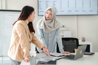 İş kıyafeti giymiş iki genç Asyalı kadının mali raporları tartışırken görüntüsü. Takım planlama için bir dizüstü bilgisayar, hesap makinesi ve veri kullanarak işbirliği yapıyor. Modern bir ofiste başarılı bir muhasebe toplantısı.
