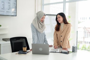 İki genç Asyalı kadın, biri başörtüsü takıyor, modern bir ofiste işbirliği yapıyor, finansal belgeleri analiz ediyor, stratejileri tartışıyor ve gülümsüyor. Takım çalışması ve işbirliği kavramı.