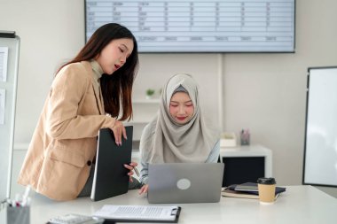 Biri tesettürlü iki genç Asyalı kadın modern bir ofiste dizüstü bilgisayarla iş birliği yapıyor. İş stratejisi, finansal planlama ve başarı için takım çalışması tartışması.