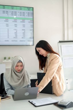 İki Asyalı kadın, biri başörtüsü takıyor, bir iş toplantısında işbirliği yapıyor, bir dizüstü bilgisayardaki mali veri analizini tartışıyor, modern bir ofiste yatırım planlama ve büyüme stratejilerine odaklanıyor.