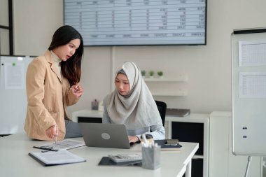 İki genç Asyalı kadın parlak bir ofiste mali raporları analiz ediyor. Bir tanesi, tesettürde, dizüstü bilgisayar kullanıyor. Takım çalışması, işbirliği ve profesyonel atmosfer görülebilir..