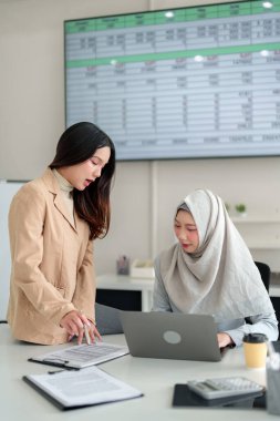 Finans belgelerini inceleyen ve modern bir ofiste dizüstü bilgisayar kullanan iki genç iş kadını (biri tesettür takıyor). İş büyümesi için takım çalışması, işbirliği ve mali analiz.