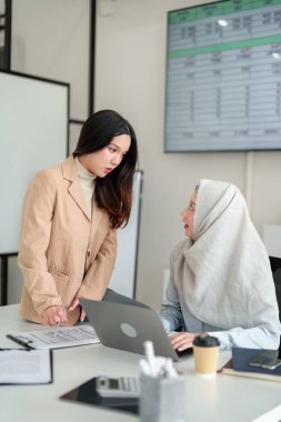 İki genç iş kadını ofiste iş birliği yapıyor. Başka bir kadın belgeleri incelerken, tesettürlü Asyalı bir kadın dizüstü bilgisayar kullanıyor. Finansal raporları ve yatırım stratejilerini tartışıyorlar..