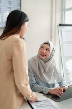 Ofiste iş birliği yapan iki genç Asyalı kadın. Biri tesettürlü, dizüstü bilgisayarla çalışıyor. Takım çalışması, iletişim ve modern Müslüman profesyonel temaları. İş yeri kavramlarında çeşitlilik için ideal.