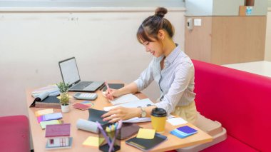 Bu görüntü, iç mimar olarak çalışan genç bir Asyalı kadını gösteriyor. Masasında oturmuş renk paletlerini, kumaş örneklerini ve mimari planlarını gözden geçirirken bir de ev yenileme projesi planlıyor..