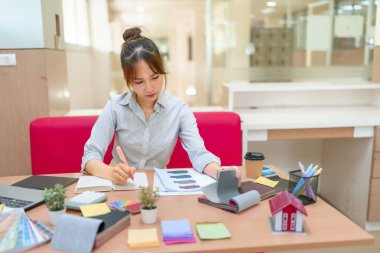 İç mimar olarak çalışan genç bir Asyalı kadın, kumaş örneklerini dikkatlice seçiyor ve ofisinde notlar alıyor. Tasarım sürecine odaklanmış durumda..