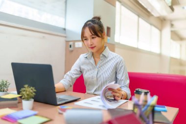 Asyalı kadın dizüstü bilgisayarına odaklanmış, evrak okuyor. Çevrimiçi kurs çalışması, araştırma kağıtları ya da tasarım projesi. Eğitim ve öğrenme kavramı. Ofis masası ortamı