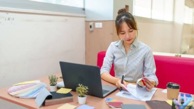 Asyalı bir iç mimar masasında dizayn ve proje detaylarını gözden geçiriyor. Renk örnekleriyle çalışıyor ve modern ofis ortamında notlar alıyor..