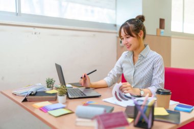 Genç bir Asyalı kadın iç mimar dizüstü bilgisayar üzerinde çalışıyor ve masasındaki renk örneklerini inceliyor. Müşterileri için yeni bir iç tasarım projesi hazırlıyor..